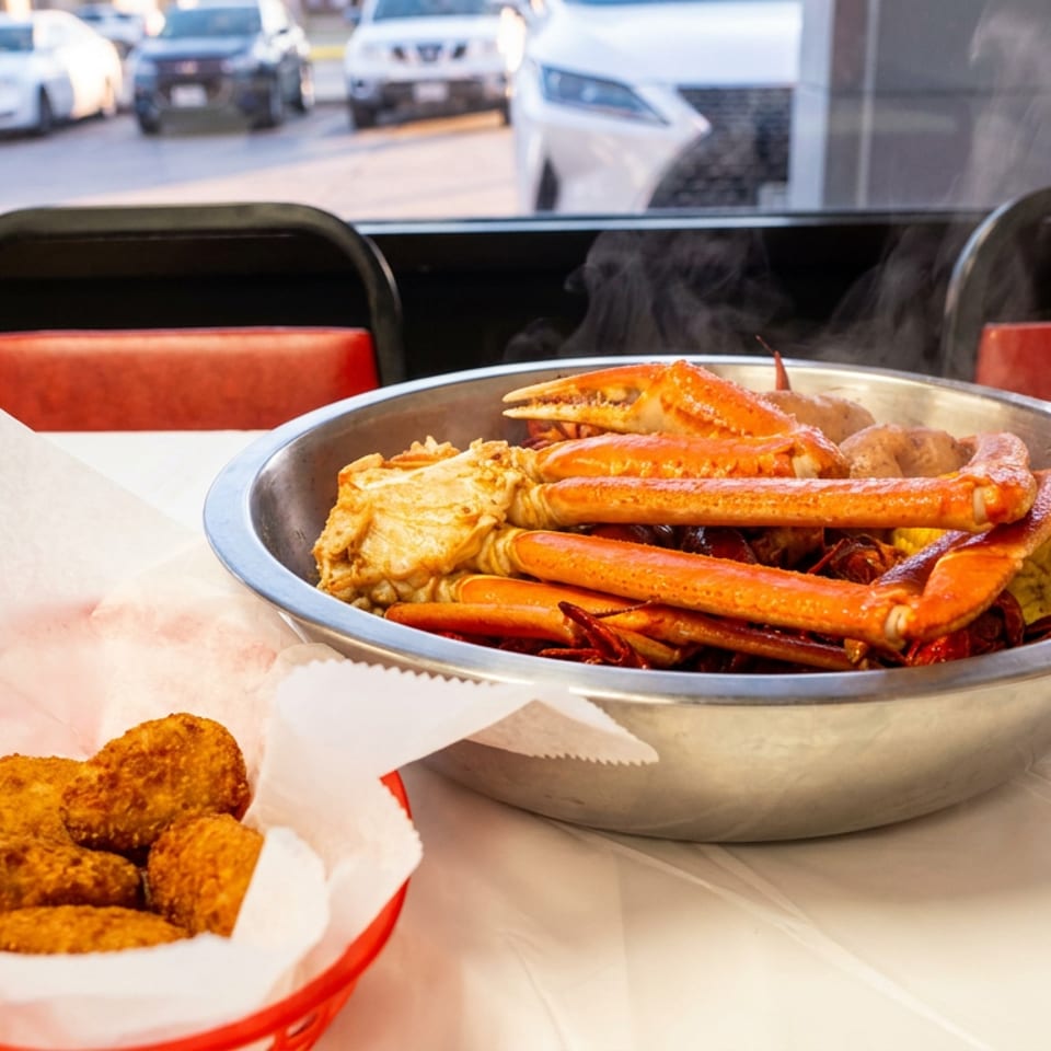 Steaming crab legs, corn, and potatoes in a metal bowl, with fried appetizers in a basket.